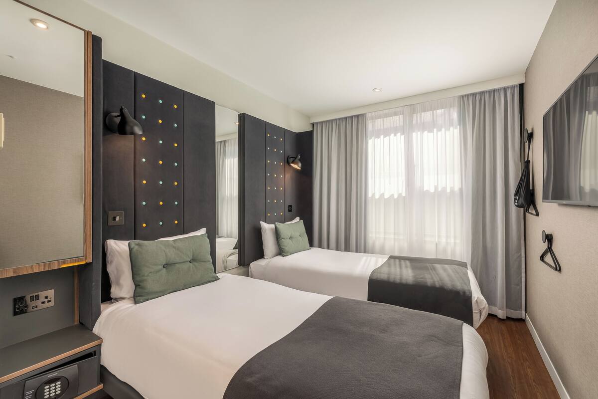 Habitación de hotel con dos camas, un espejo grande, un armario con iluminación y una ventana con cortinas.