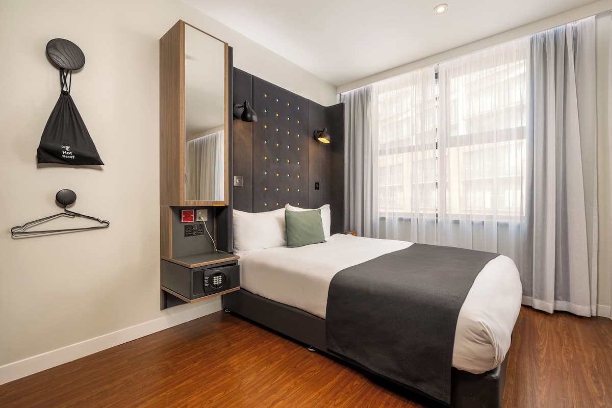 Una habitación de hotel con cama, mesita de noche, espejo, bolso colgado y ventana con cortinas.