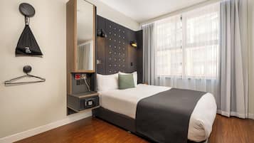 Une chambre d’hôtel avec un lit, une table de chevet, un miroir, un porte-manteau et une fenêtre avec des rideaux.