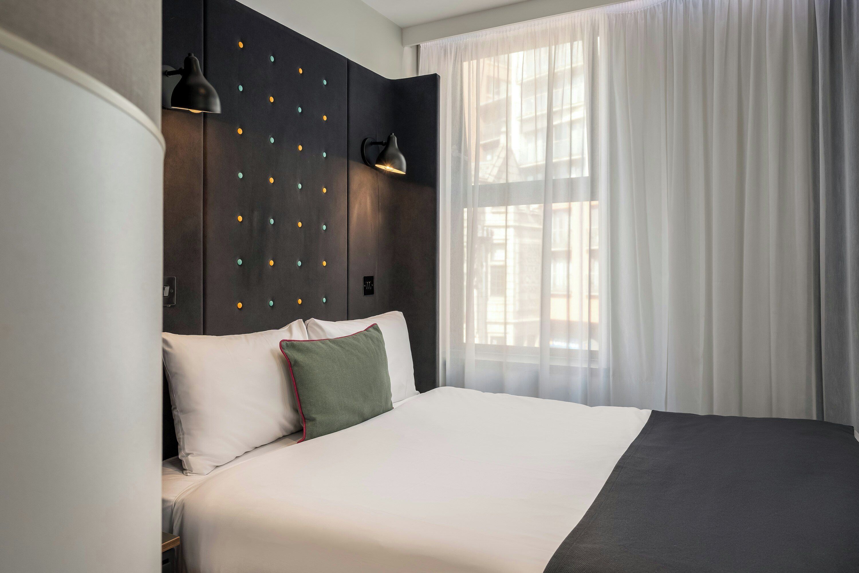 A modern hotel room with a large bed, a dark headboard with decorative lights, a bedside lamp, and sheer curtains.