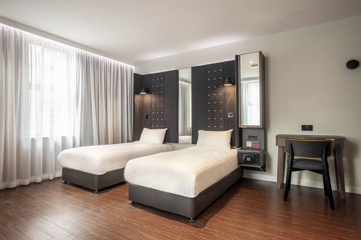 Habitación de hotel con dos camas, un escritorio, una silla, un espejo grande y una mesita de noche con lámpara.