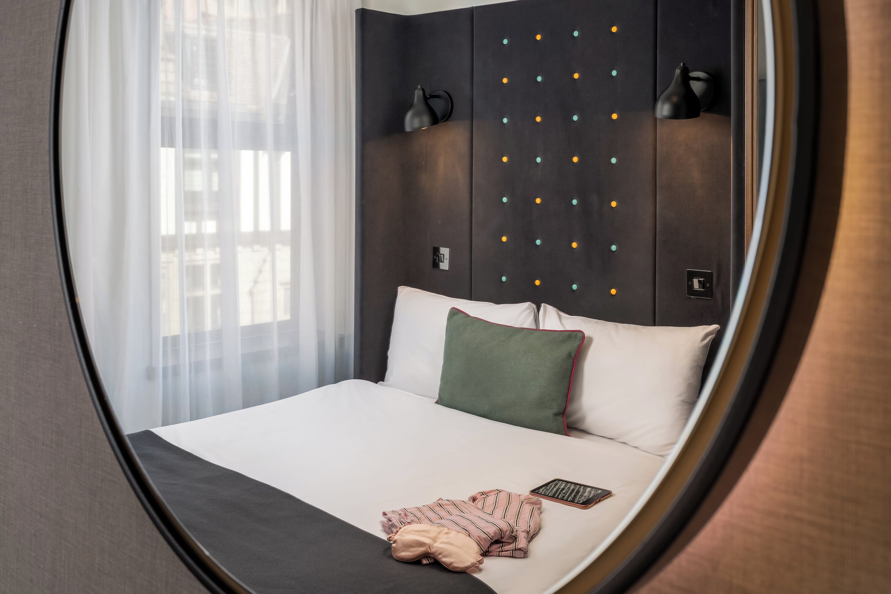 A hotel room with a bed, a bedside table, a lamp, a window with sheer curtains, and a wall with a decorative light fixture.