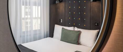 A hotel room with a bed, a bedside table, a lamp, a window with sheer curtains, and a wall with a decorative light fixture.