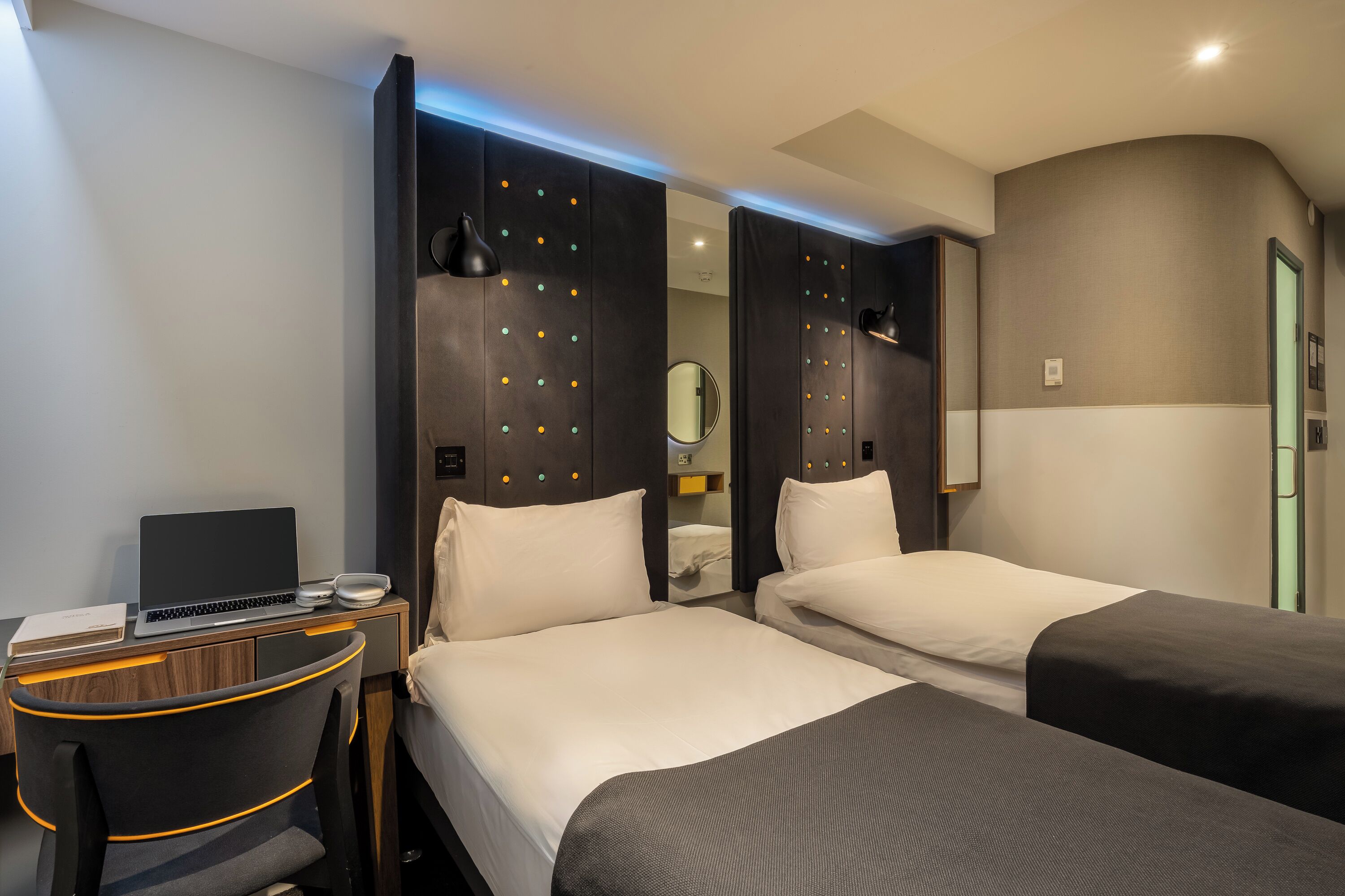 Hotellihuone, jossa on kaksi sänkyä, työpöytä, jossa on kannettava tietokone, tuoli ja peili.