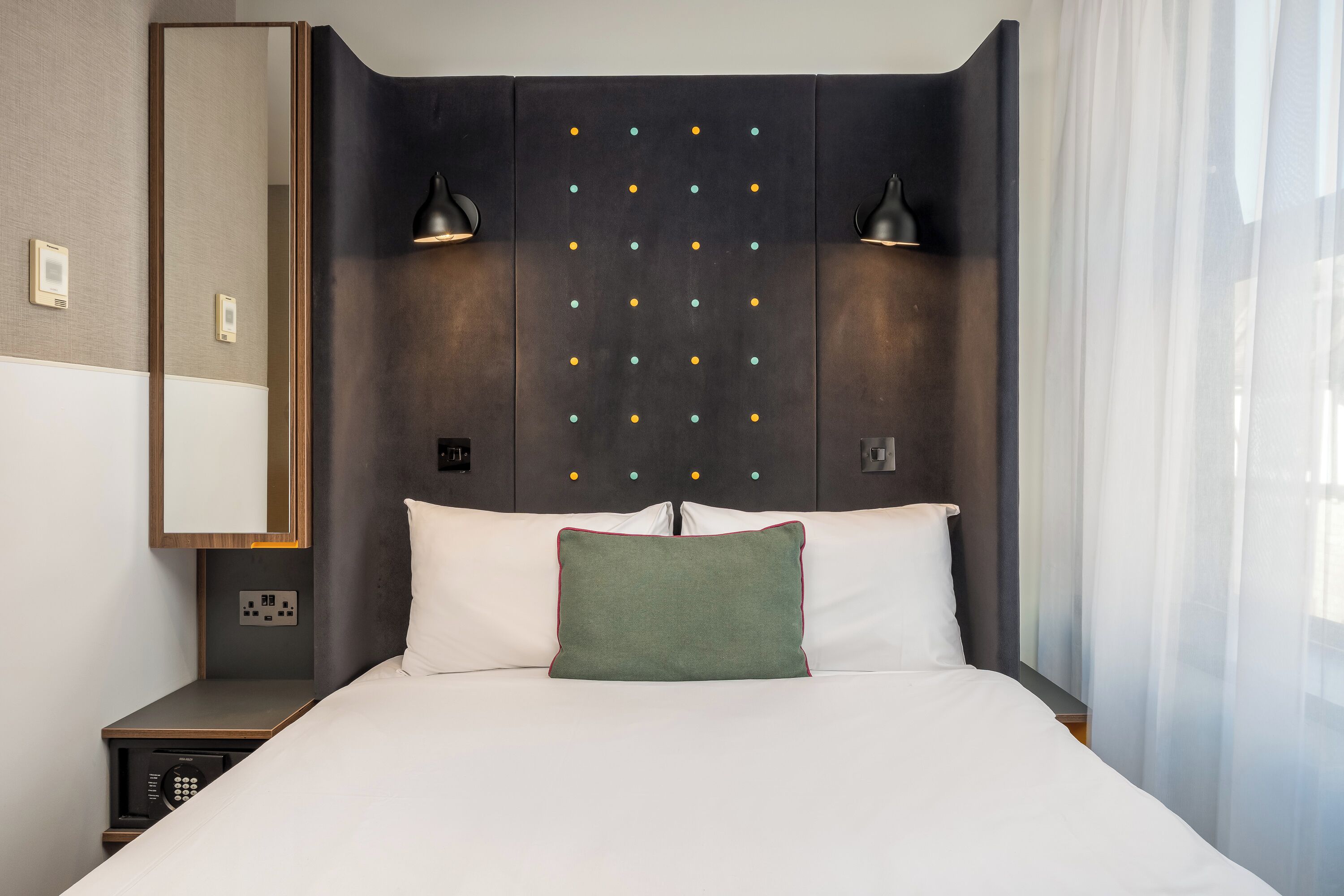 A hotel room with a bed, a nightstand with a phone, a mirror, and a wall with a grid of lights.