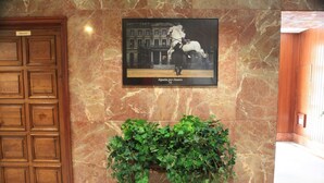 A lobby with a wooden door, a framed photograph of a horse, and a green plant on a stand. - Hotel El Coloso (Jerez de la Frontera)