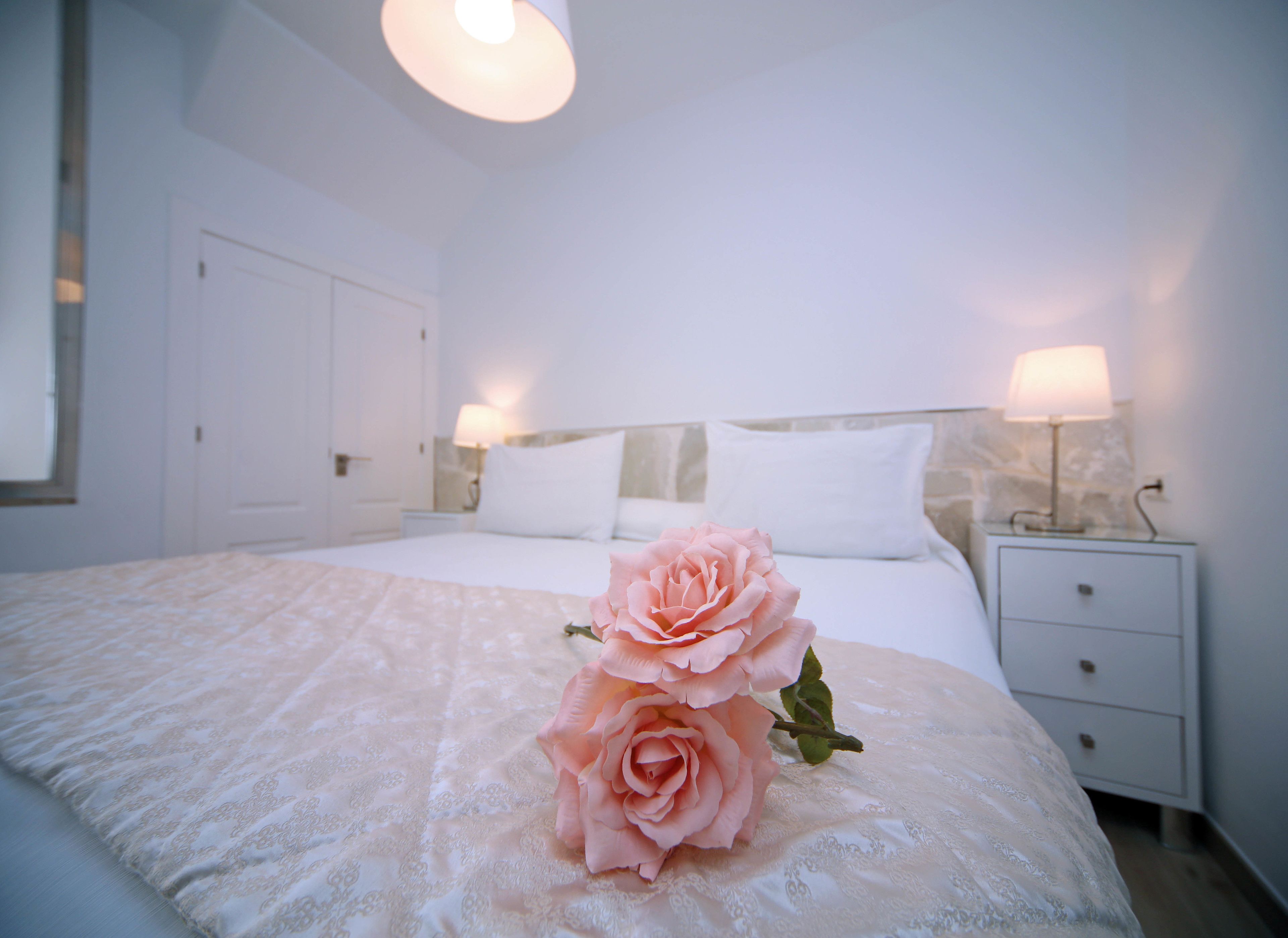 Une chambre à coucher avec un grand lit, du linge de lit blanc, deux lampes de chevet et une seule rose rose posée sur le lit.