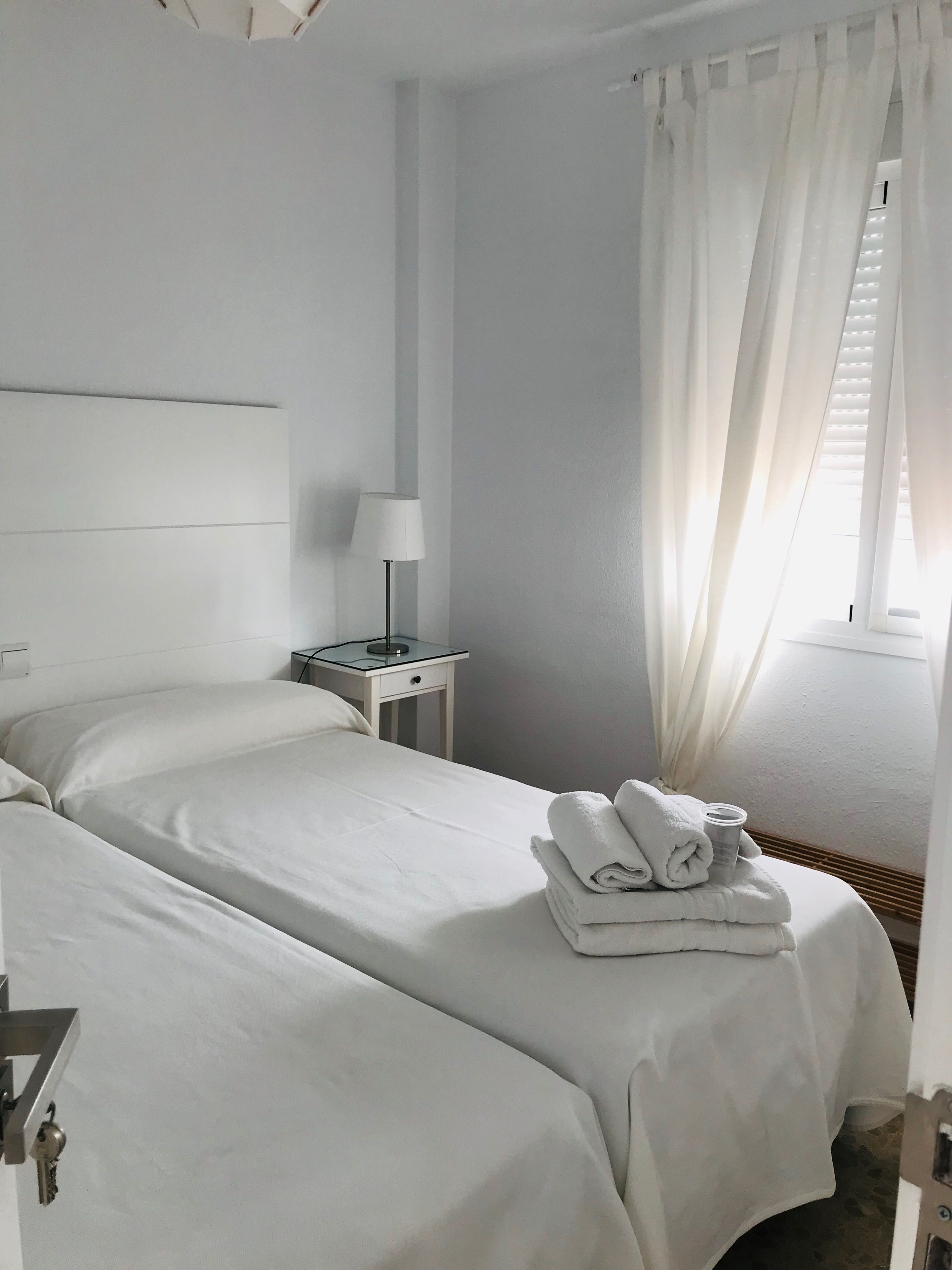 A hotel room with two beds, a bedside table with a lamp, and a window with sheer curtains.