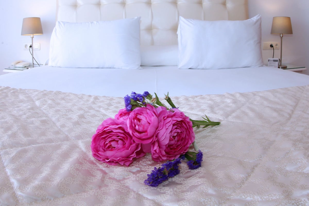 A hotel room with a tufted headboard, white bedding, two bedside lamps, and a bouquet of pink and purple flowers on the bed.