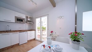 Una cocina moderna con armarios blancos, un microondas y un lavavajillas. Hay una mesa de comedor con una bandeja de tazas blancas y una planta en maceta con flores rosas.