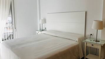 A neatly made bed with white linens, a bedside table with a lamp, and a window with curtains.
