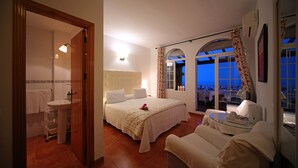 Un dormitorio con cama, espacio para sentarse y vista a la ciudad al atardecer.