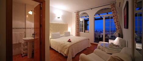 A bedroom with a bed, a sitting area, and a view of the city at dusk.