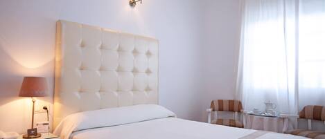 Een hotelkamer met een groot bed, een nachtkastje met een lamp, een klein tafeltje met een theepot, en een stoel.