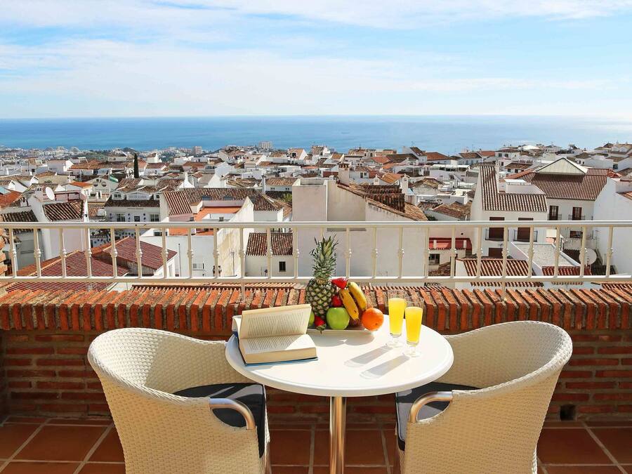 Una terraza con dos sillas de mimbre, una mesa, un frutero y un vaso de zumo de naranja.