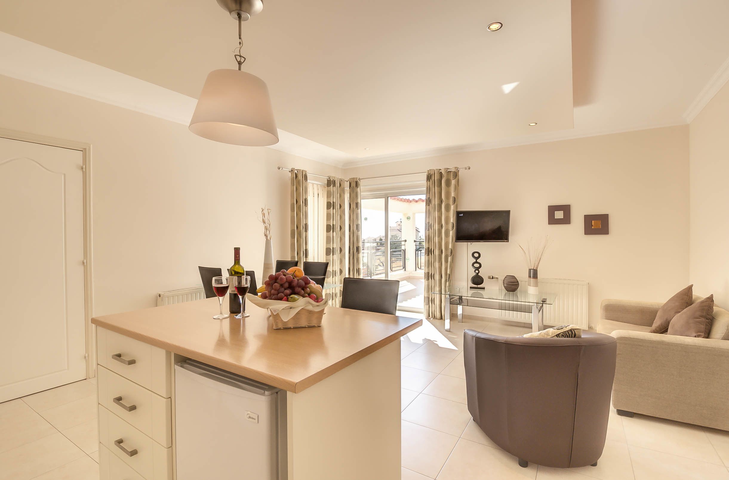 A modern kitchen with a central island, a dining area with a glass table, and a living room with a sofa and TV.