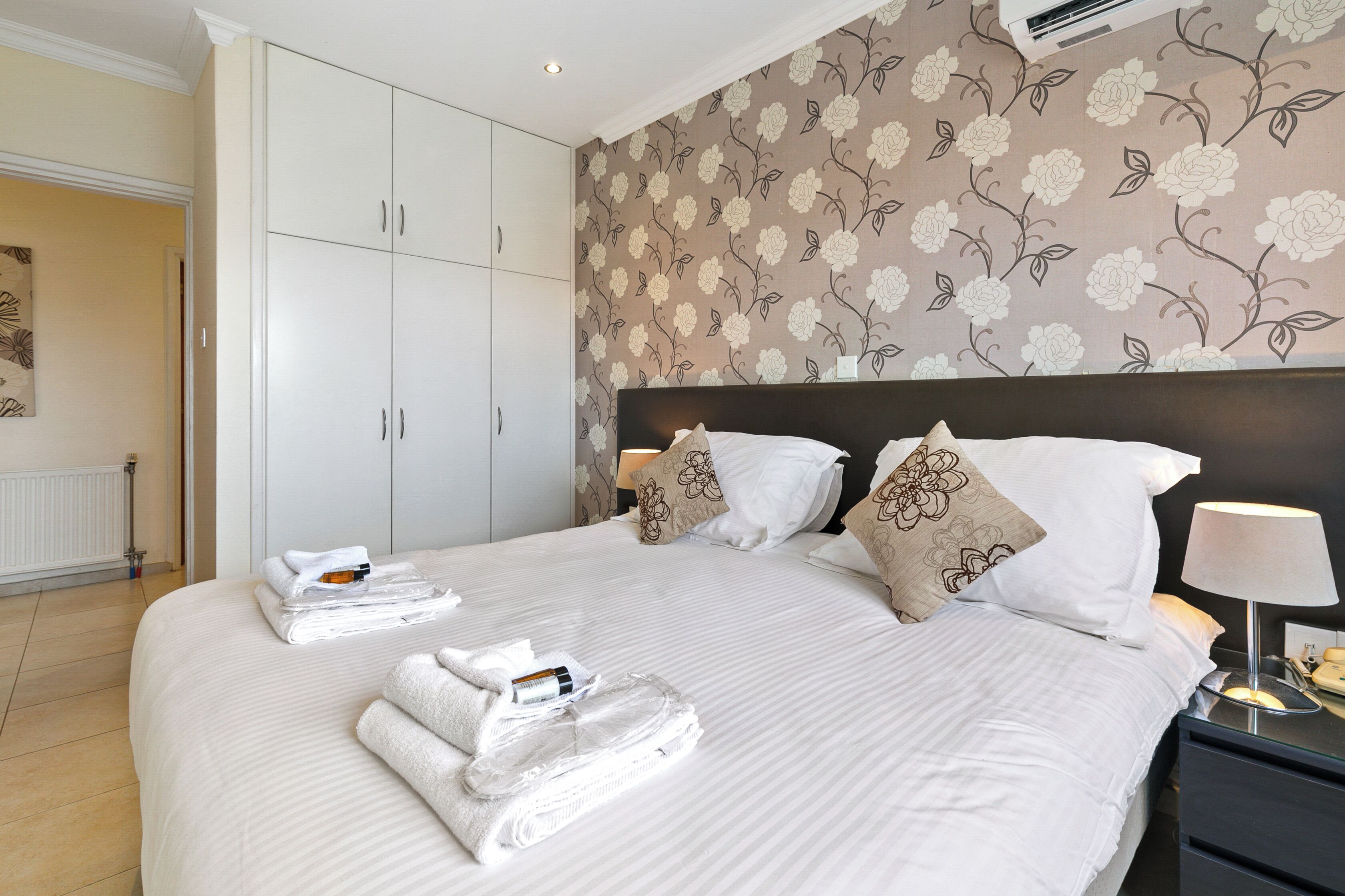 A hotel room with a large bed, white bedding, a floral wallpaper, and a nightstand with a lamp.