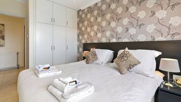 A hotel room with a large bed, white bedding, a floral wallpaper, and a nightstand with a lamp.