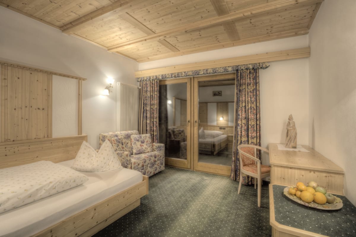 A cozy room with a wooden ceiling, a bed with patterned pillows, a small table with fruit, and a chair.