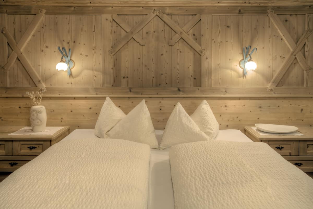A double bed with white linens and three pillows, wooden headboard with a cross-brace design, and two wall-mounted wall sconces.