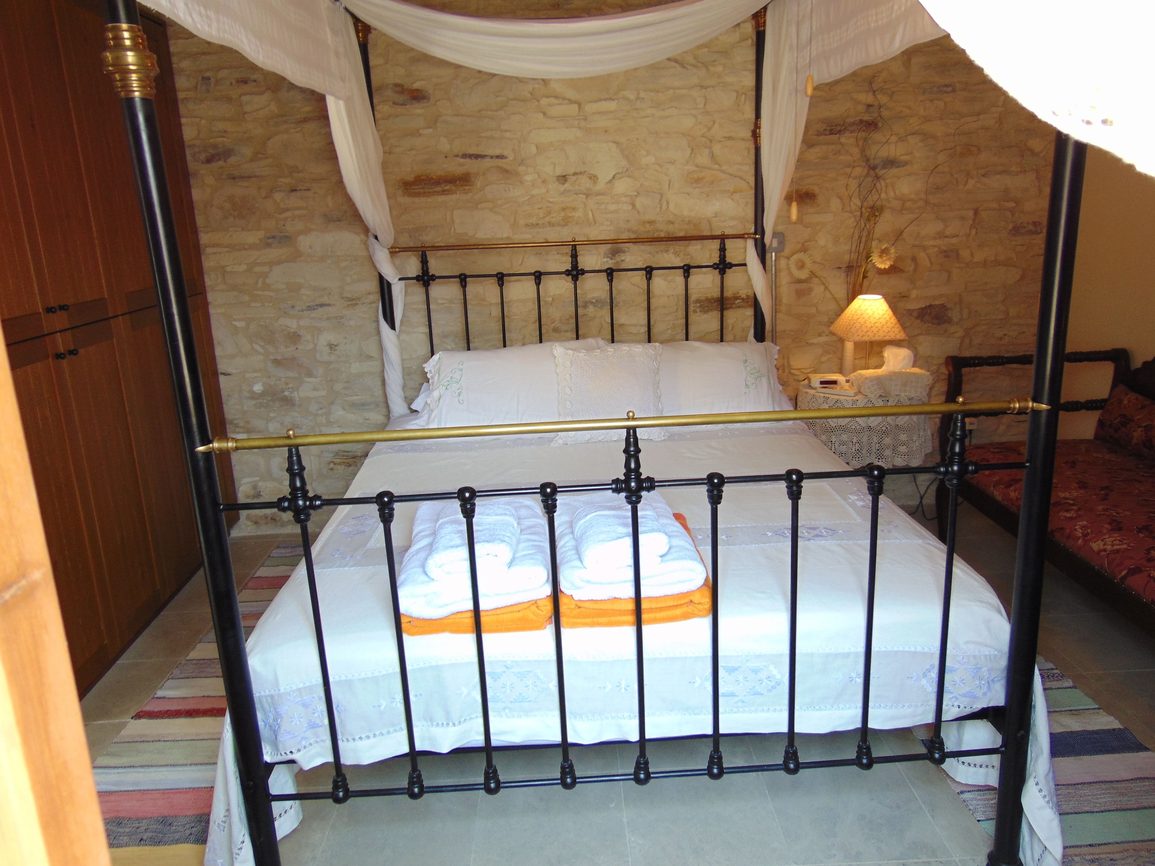Una cama con dosel sobre cuatro columnas, una pared de piedra y una mesita de noche con una lámpara.