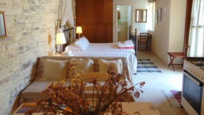 Een slaapkamer met een stenen muur, een houten plafond, een bed met witte lakens, een nachtkastje met lampen, een stoel, een kleine tafel met bloemen, en een keuken met een fornuis.