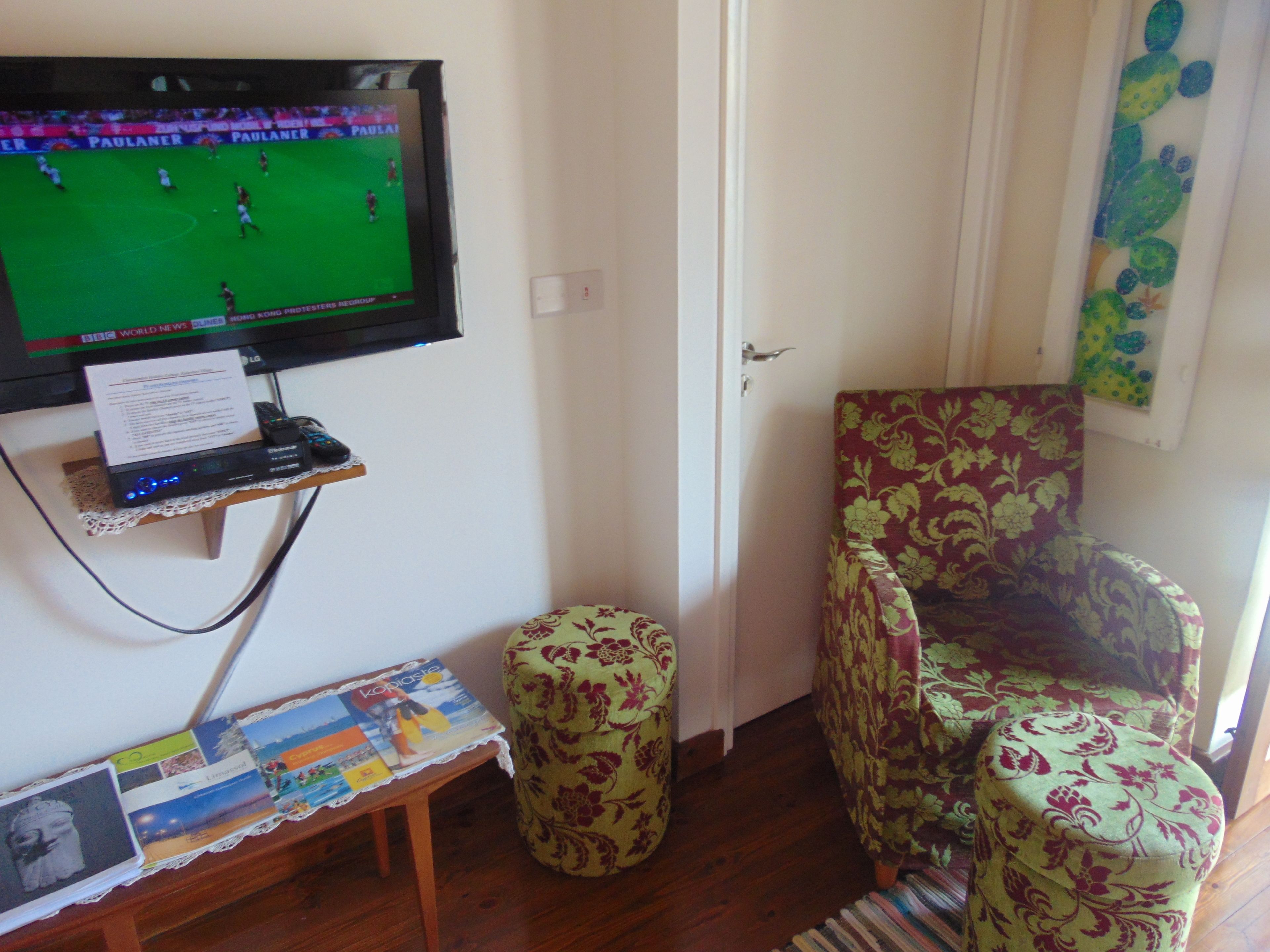A living room with a television displaying a sports game, a small table with magazines, a patterned armchair, and a wooden floor.