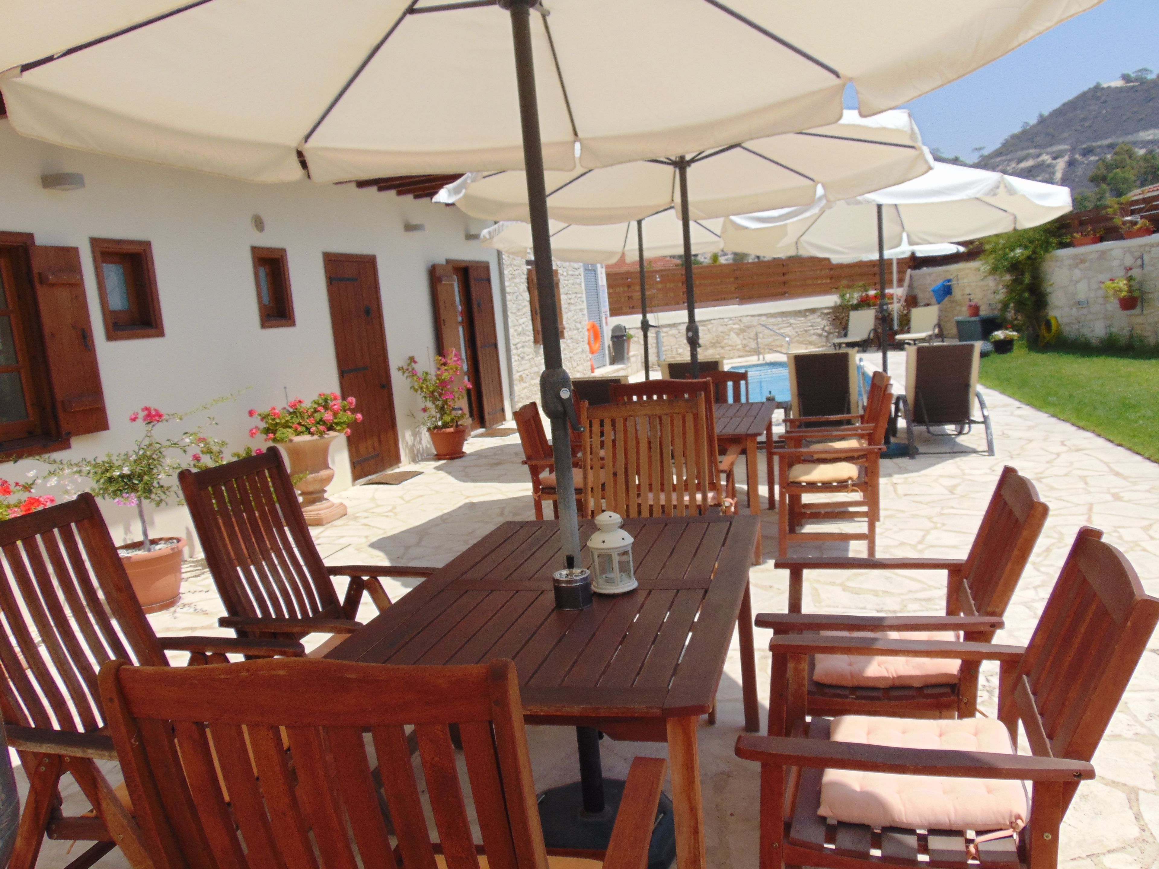 An outdoor patio with wooden tables and chairs, umbrellas, potted plants, and a stone wall.