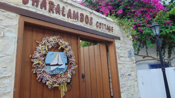 Een stenen muur met een houten deur en een bordje met de tekst 'CHARALAMOOS COTTAGE'.