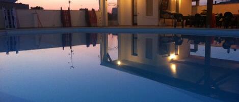 Ein Swimmingpool mit überdachtem Bereich und Liegestühlen, der die Umgebung spiegelt.