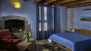 Une chambre avec une cheminée en pierre, un lit recouvert d’une courtepointe à motifs floraux bleus, une chaise en bois, une petite table avec un vase et un rideau bleu.