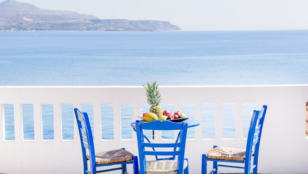 A balcony with two blue chairs and a table with fruit, overlooking a calm sea and distant hills. - Kalyves Bay (Apokoronas)