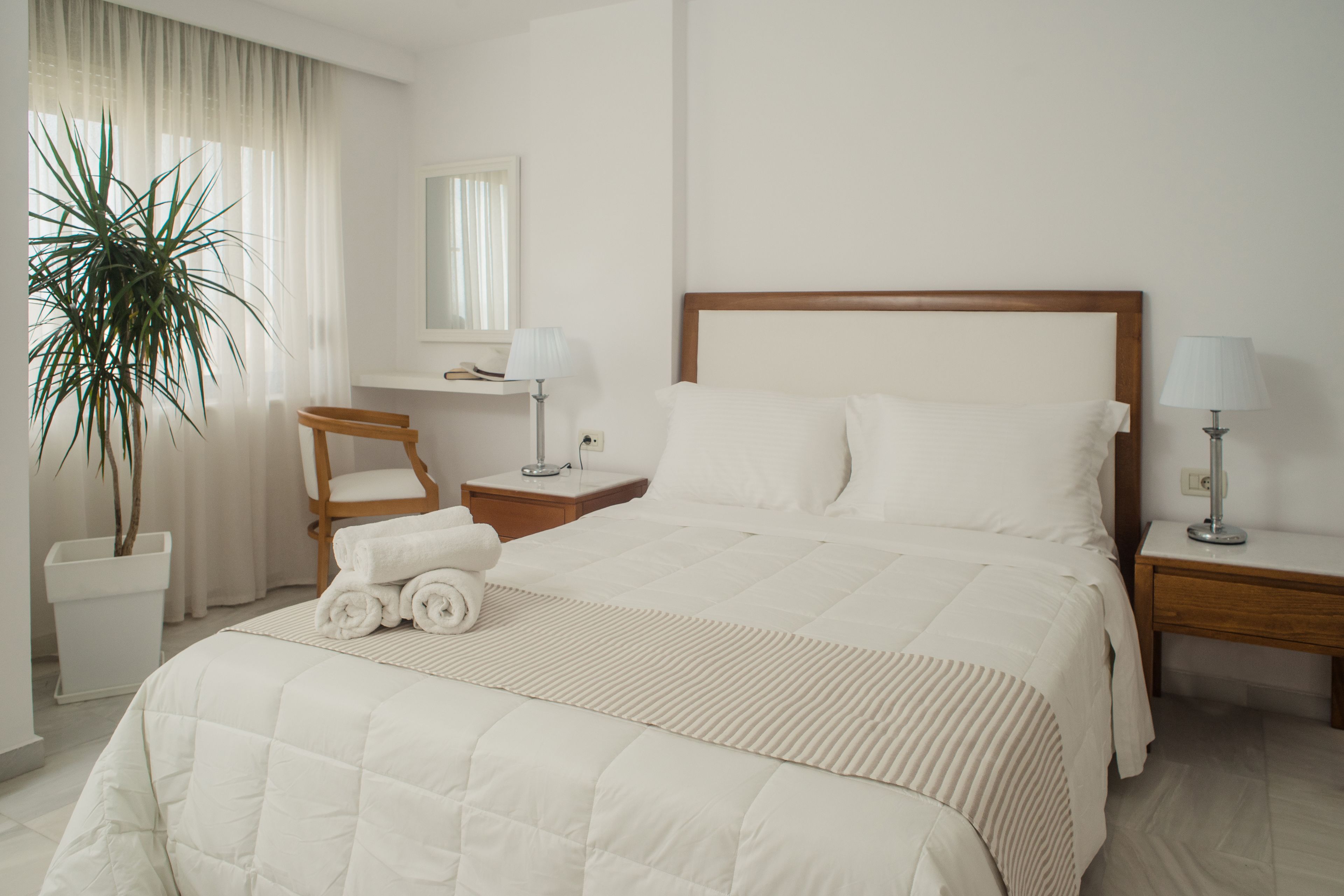 Une chambre d’hôtel avec un grand lit, une table de chevet avec une lampe, une chaise, une plante en pot et un miroir au mur.