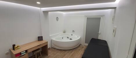 A bedroom with a wooden desk, a black massage table, a white bathtub, and a door leading to another room.