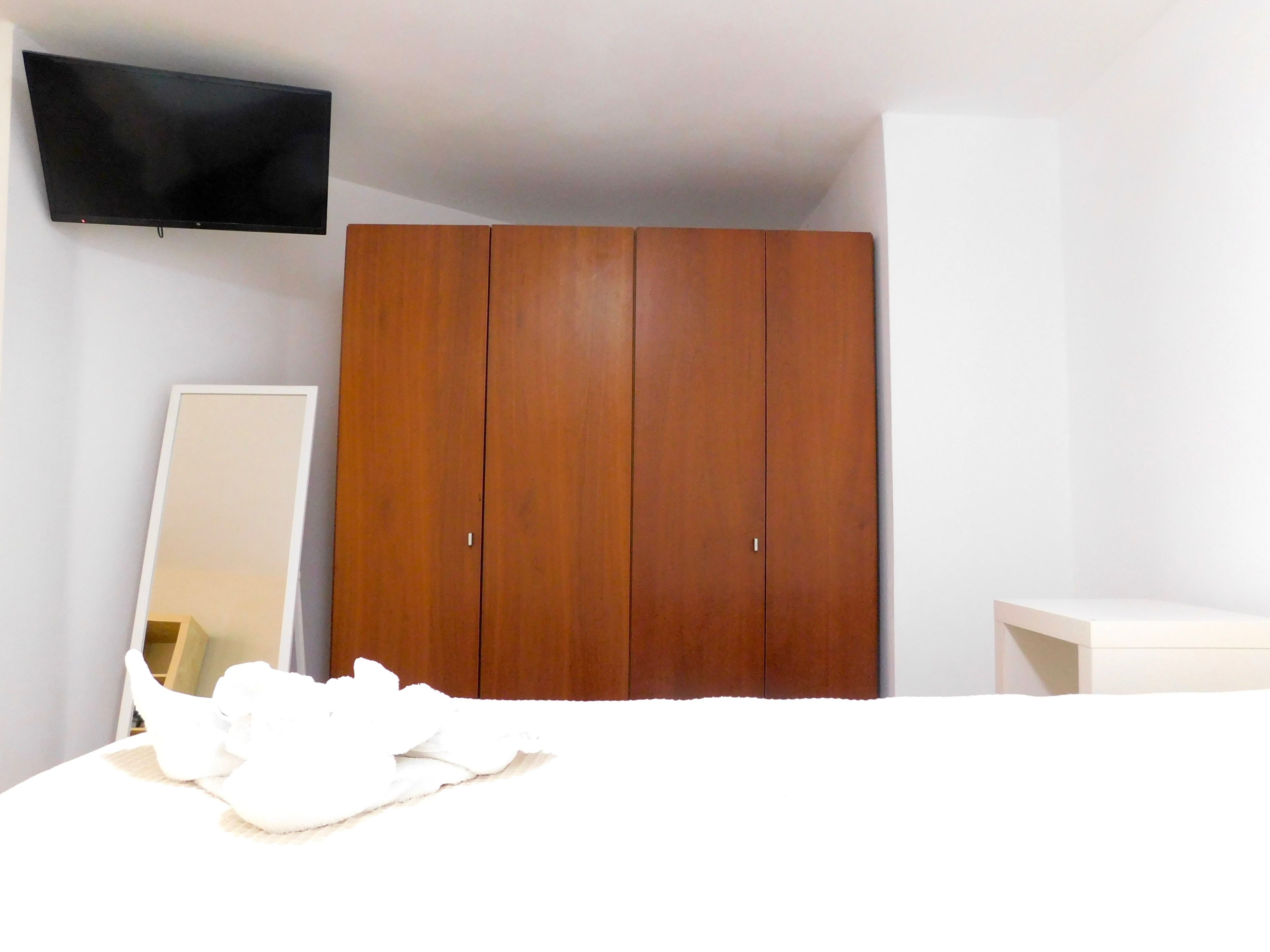 Quarto com cama, guarda-roupa de madeira, televisão na parede e mesa de cabeceira.