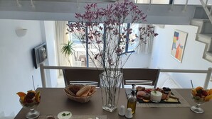 A dining table with a variety of food items, including bread, cheese, and fruits, set in a modern room with a view of the outside. - Elizabeth Suites (Chania)