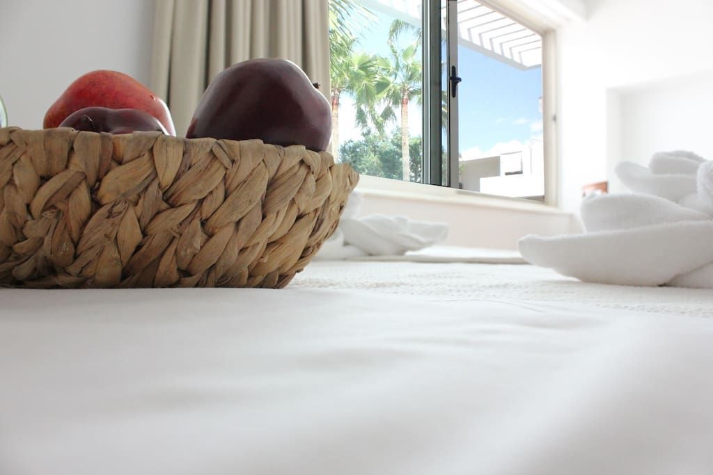 Ein Bett mit einem gewebten Korb voller Früchte, ein Fenster mit Blick auf Palmen und ein weißes Handtuch auf dem Bett.