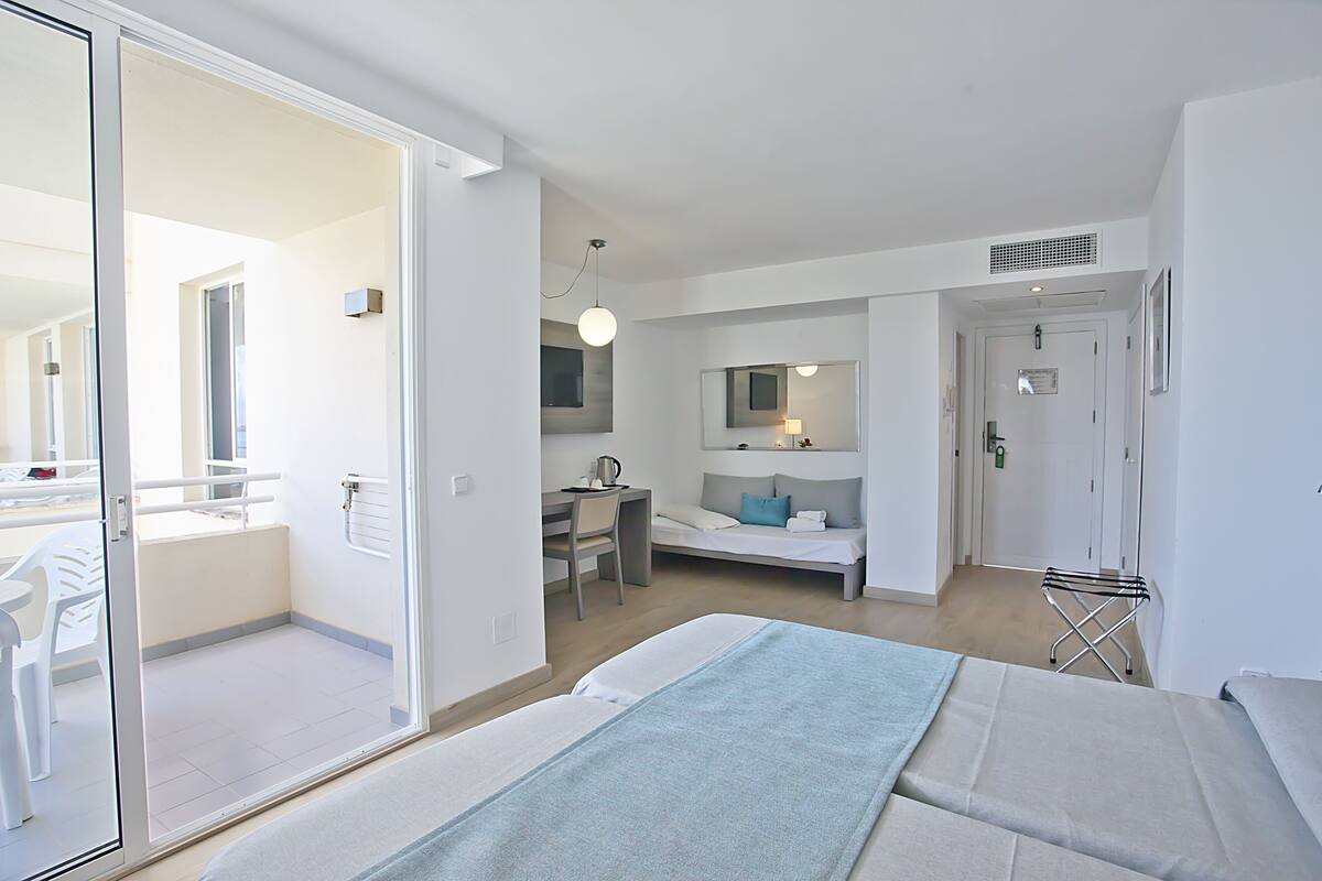 Ein modernes Hotelzimmer mit Balkon, Schreibtisch und Stuhl, einem Bett mit blauer Decke sowie einem Badezimmer mit Spiegel und Waschbecken.