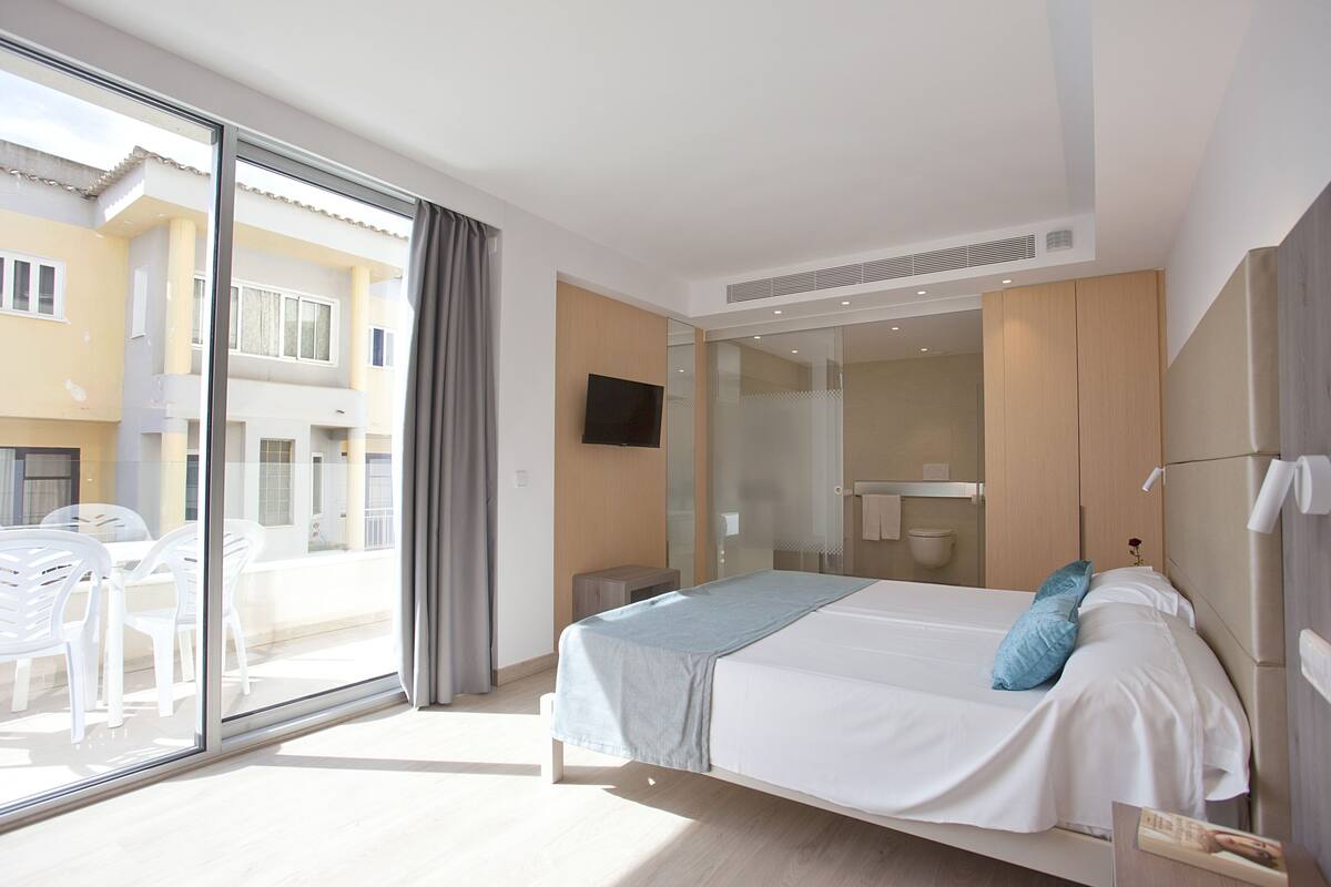 Ein modernes Hotelzimmer mit einem großen Bett, einem Fernseher, einer Glastür zu einem Außenbereich mit Stühlen und Blick auf die Außenfassade des Gebäudes.