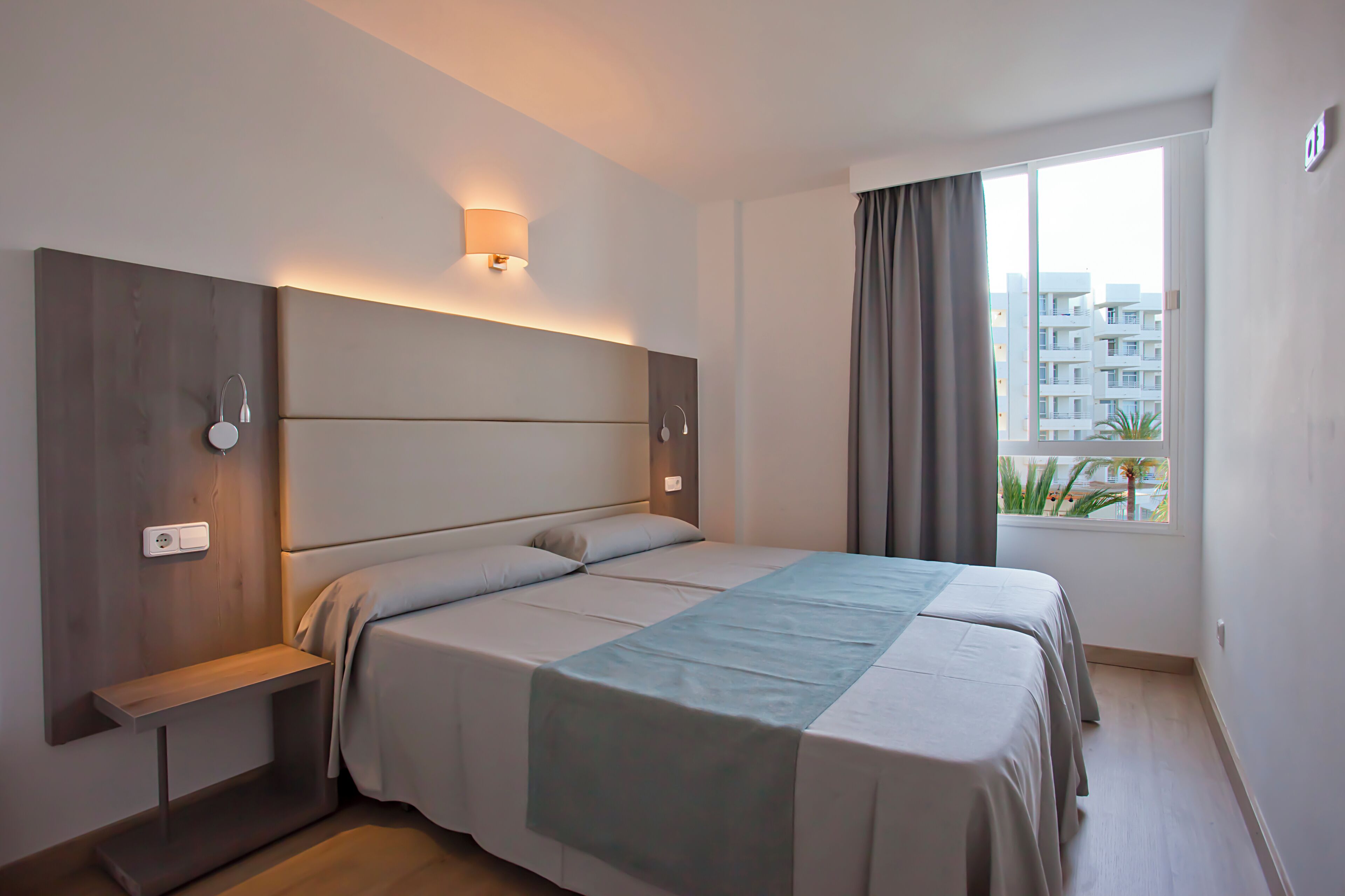 A modern hotel room with a large bed, a bedside table, a wall-mounted light, and a window with curtains.
