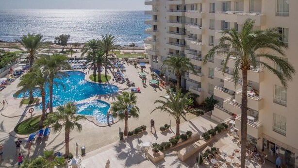 A hotel with a pool area, palm trees, and a view of the ocean. - Aparthotel Playa Dorada (Sant Llorenc des Cardassar)