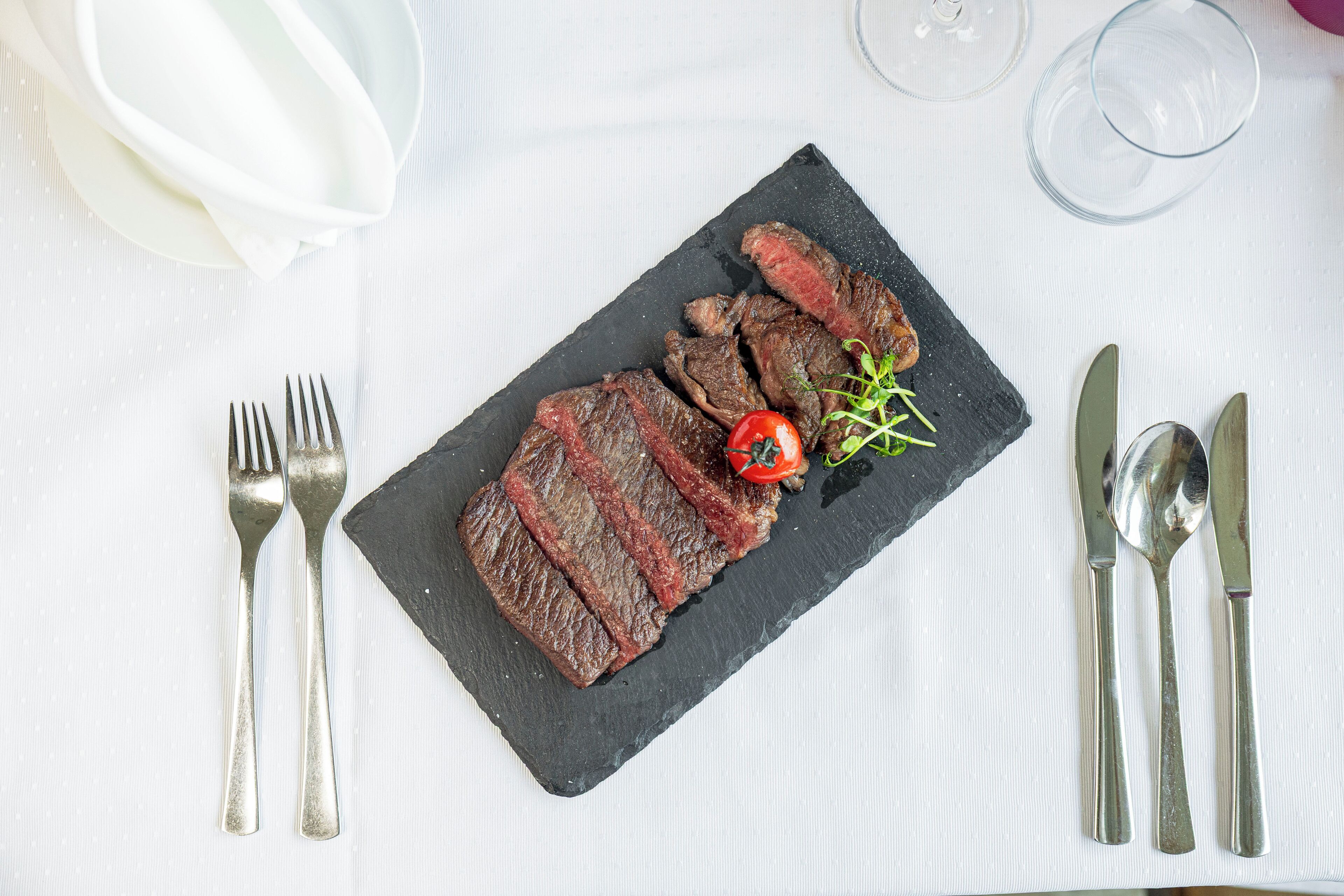 Une assiette en ardoise noire sur laquelle on a disposé des tranches de steak, une tomate cerise et une petite branche de verdure, le tout posé sur une nappe blanche.