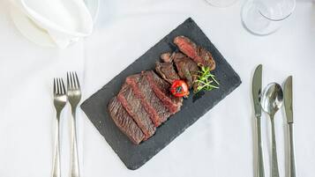A black slate plate with sliced steak, a cherry tomato, and a sprig of greenery on a white tablecloth.