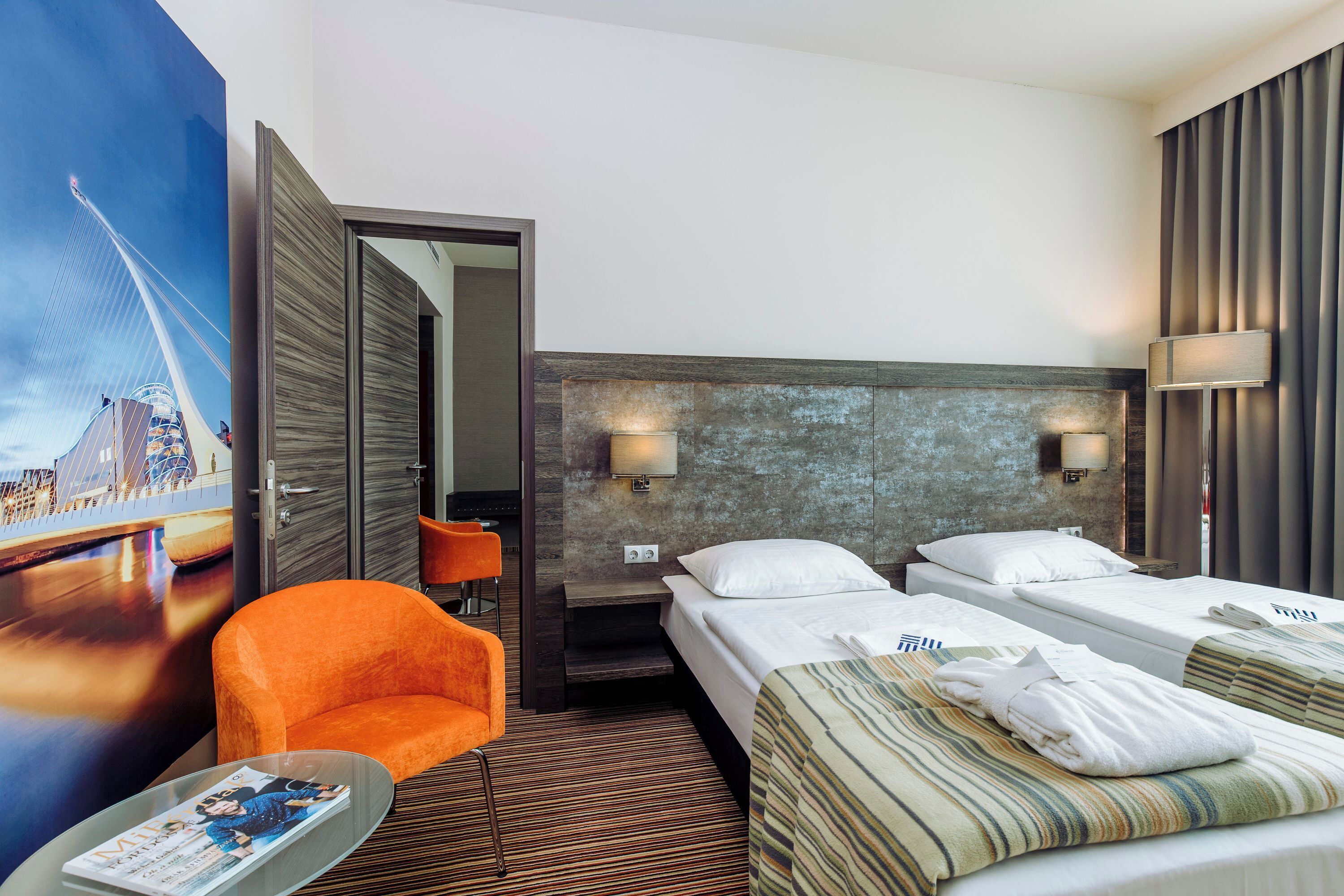 Quarto de hotel com uma cama, duas mesas de cabeceira, uma cadeira, uma televisão e um cartaz grande na parede.