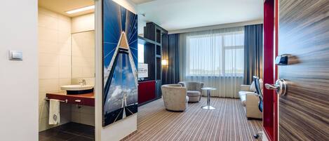 A modern hotel room with a bathroom, a seating area, and a large window.