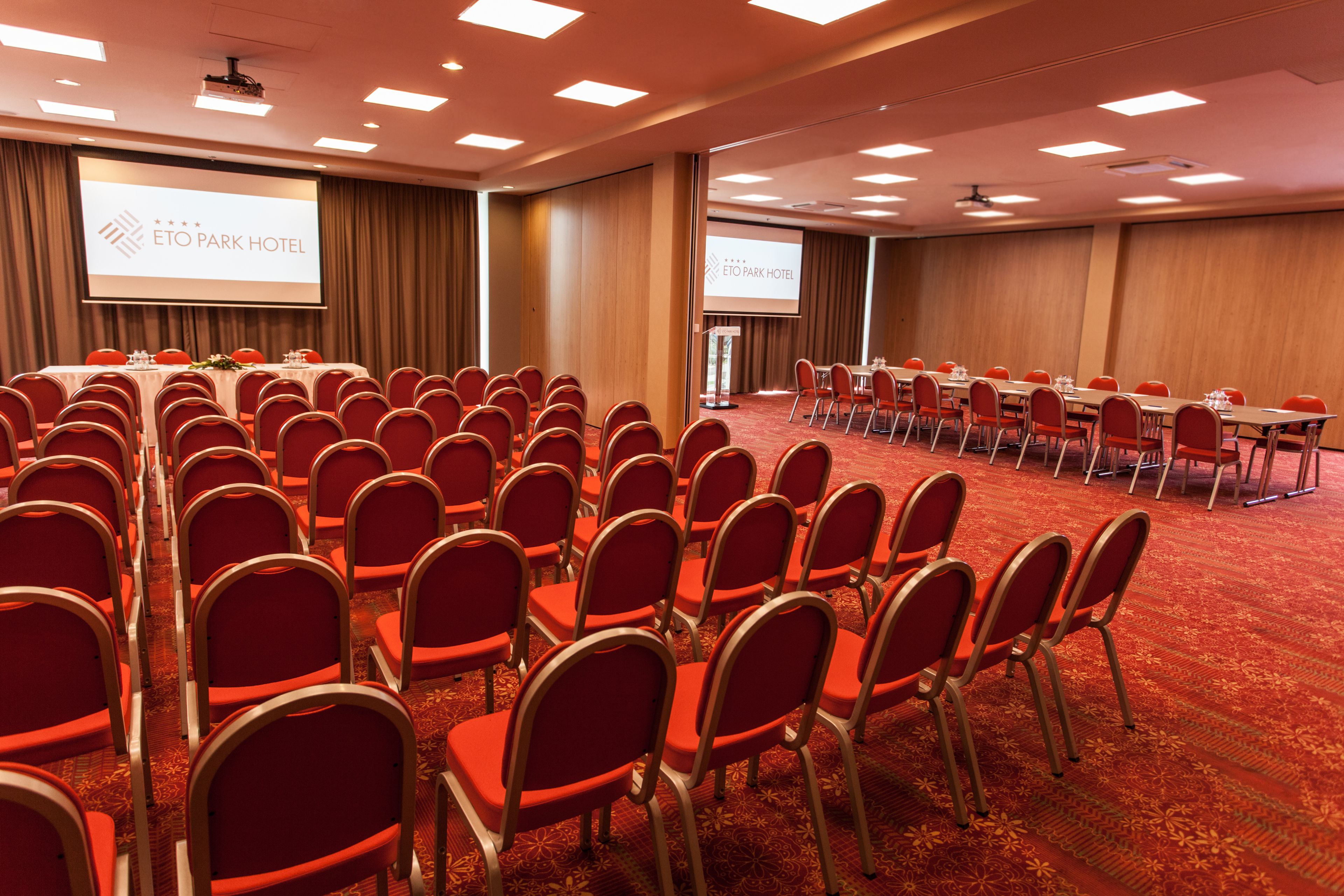 A conference room with rows of red chairs, a large screen, and a carpeted floor.