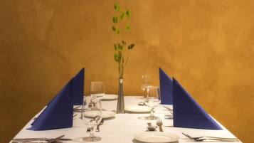 Una mesa para dos con mantel blanco, servilletas azules y un florero con hojas verdes.