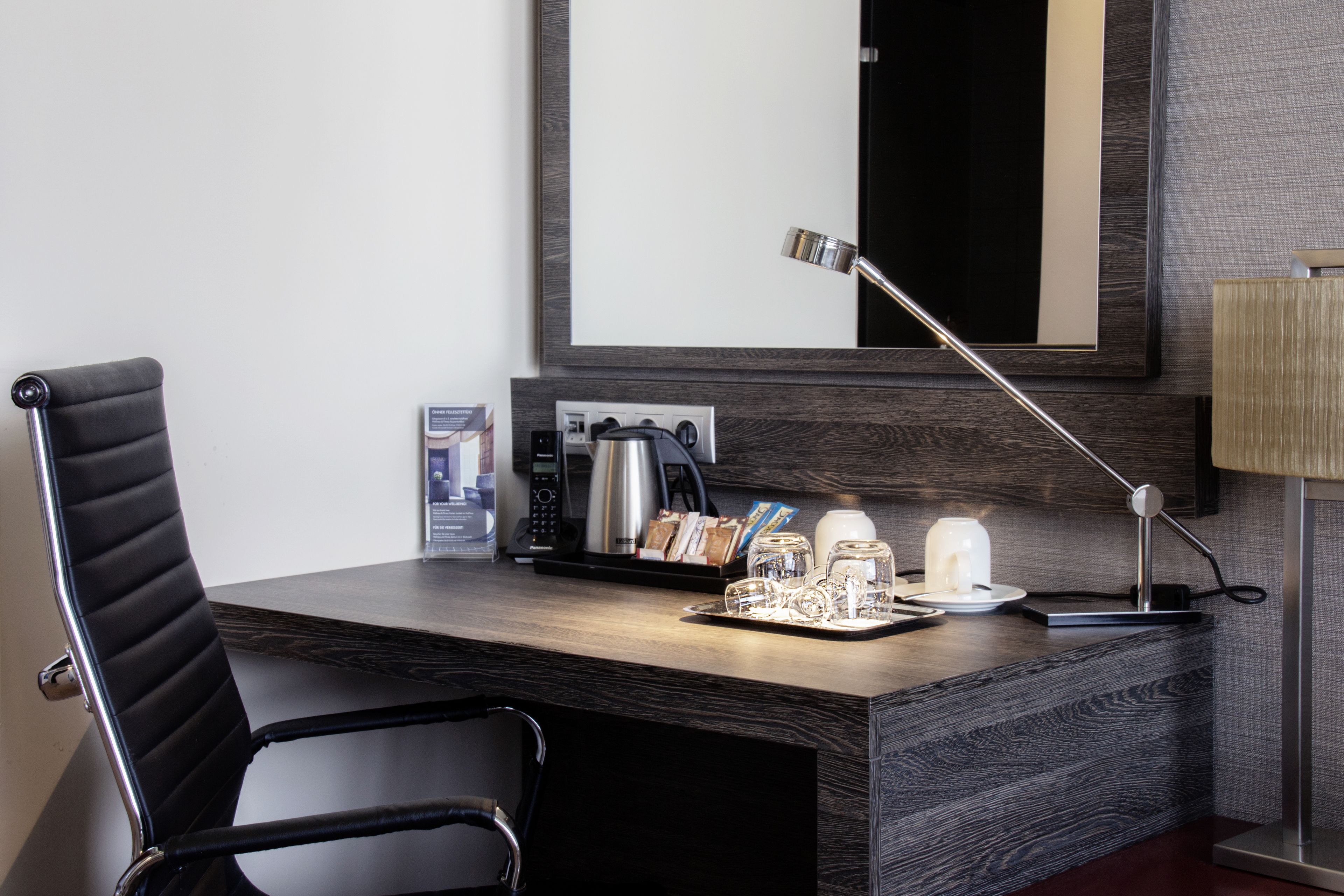 A modern hotel room with a wooden desk, a black leather chair, a coffee maker, a tray with glasses, and a lamp.