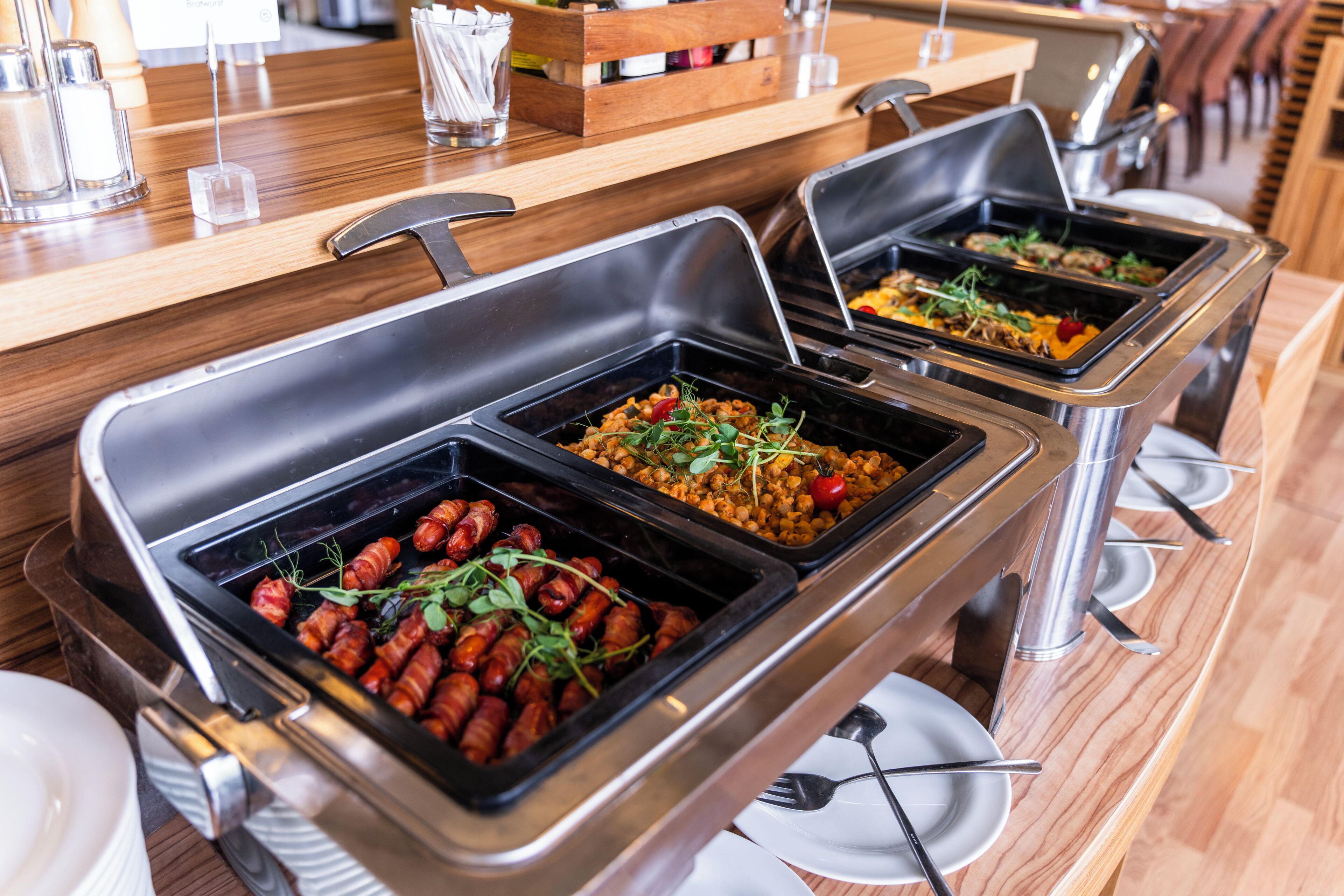 Buffet mit verschiedenen Speisen, darunter Speckröllchen und Salate, in einem modernen Ambiente mit Holztischen und Metalltabletts.