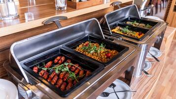 Buffet mit verschiedenen Speisen, darunter Speckröllchen und Salate, in einem modernen Ambiente mit Holztischen und Metalltabletts.